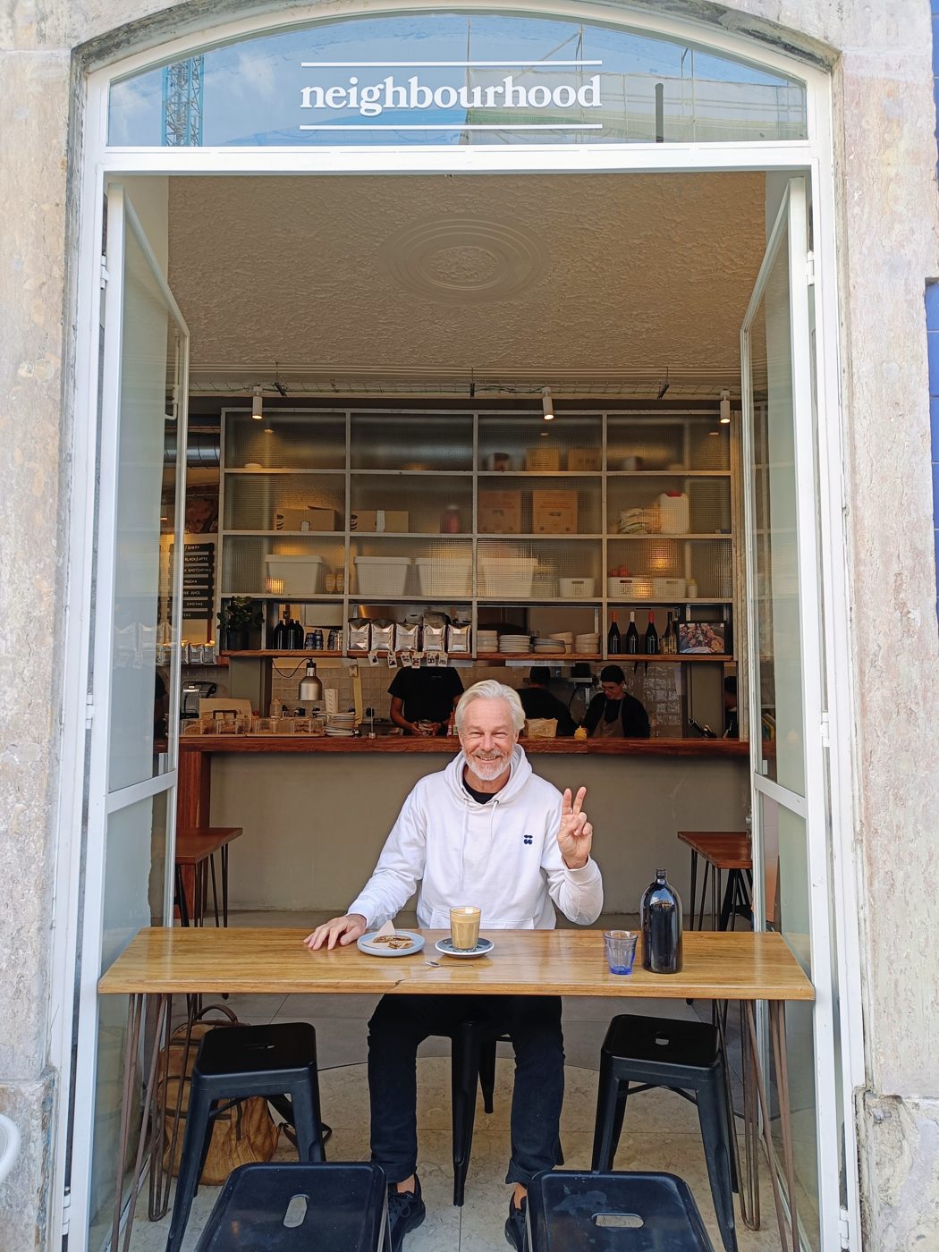 Hans at a wooden counter in the open doorway of a café called neighbourhood, giving a peace sign