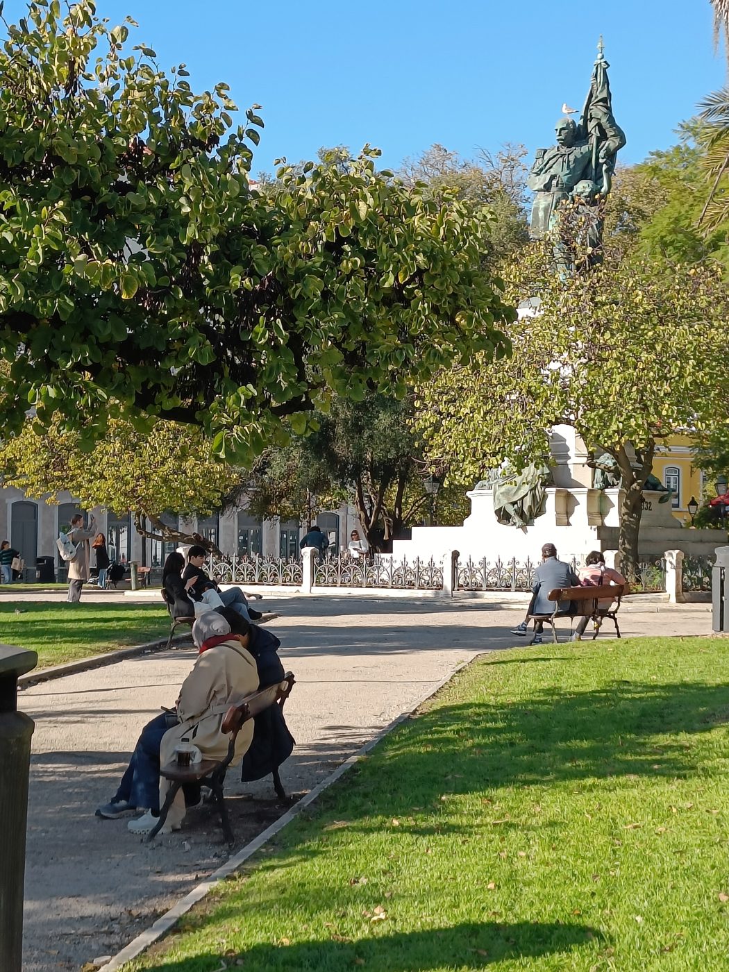 People sitting on benches in a sunny square with trees, lawn and a statue on a tall column