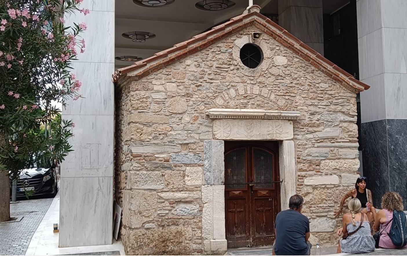 A small stone chapel between marble office columns in central Athens, people sitting on its step