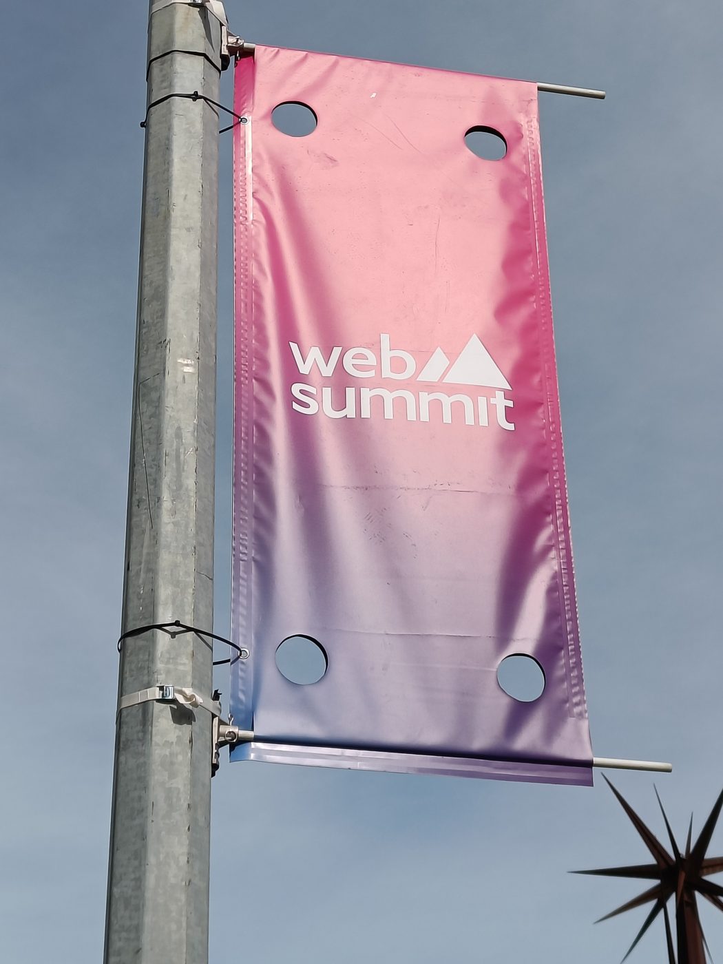 A pink Web Summit banner hanging from a lamp post against a blue sky