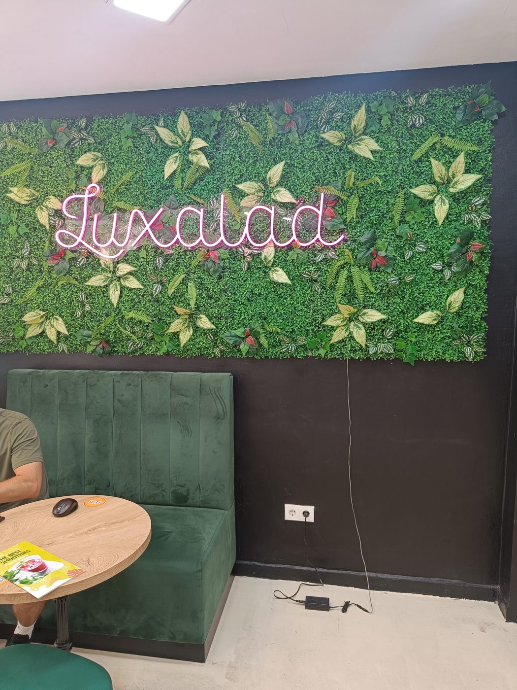 A pink Luxalad sign on a leafy café wall above green seating